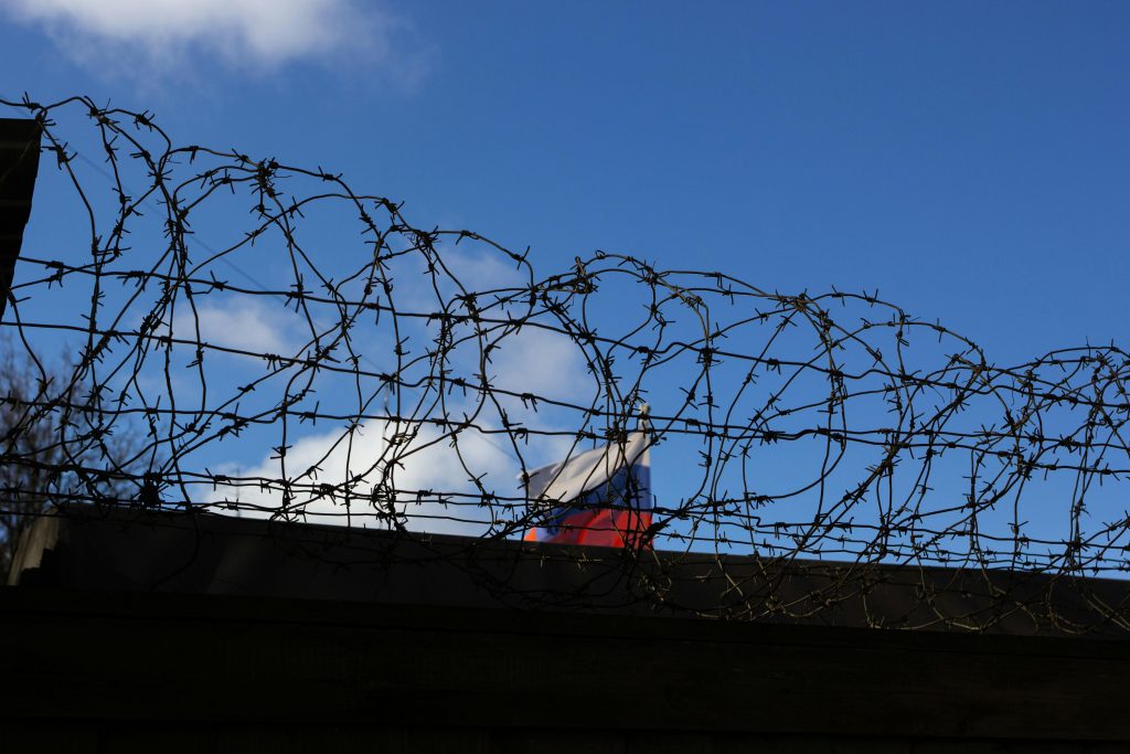 Silhouette of barbed wire with a flag in the background against a clear blue sky.