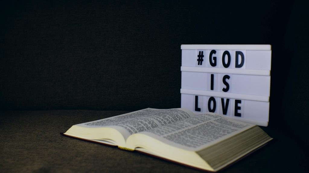 BUT FOR GOD An open Bible next to a lightbox sign displaying the text 'God is Love' on a dark background.