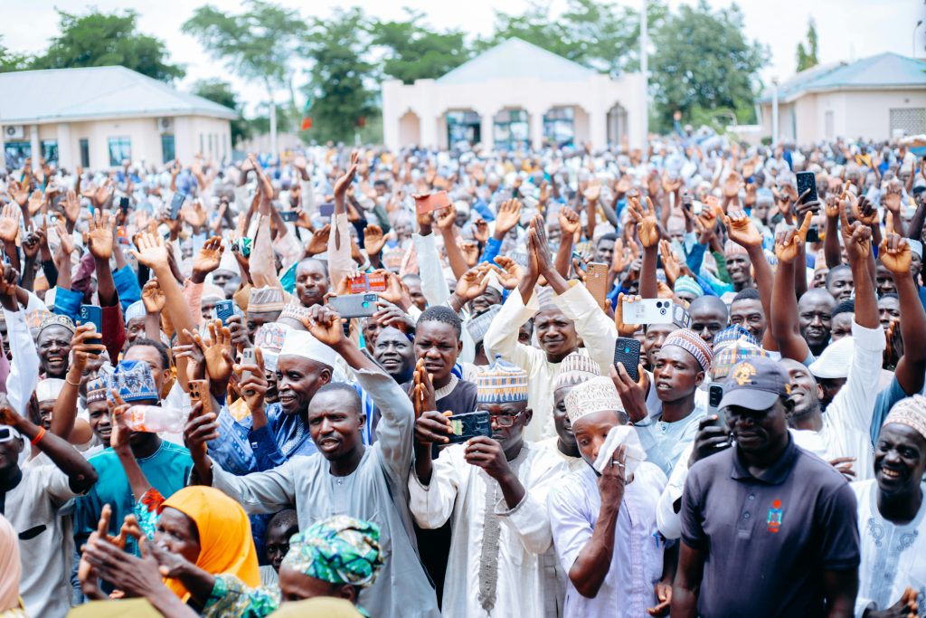 A large, lively crowd of people celebrating outdoors with raised hands and smiles.